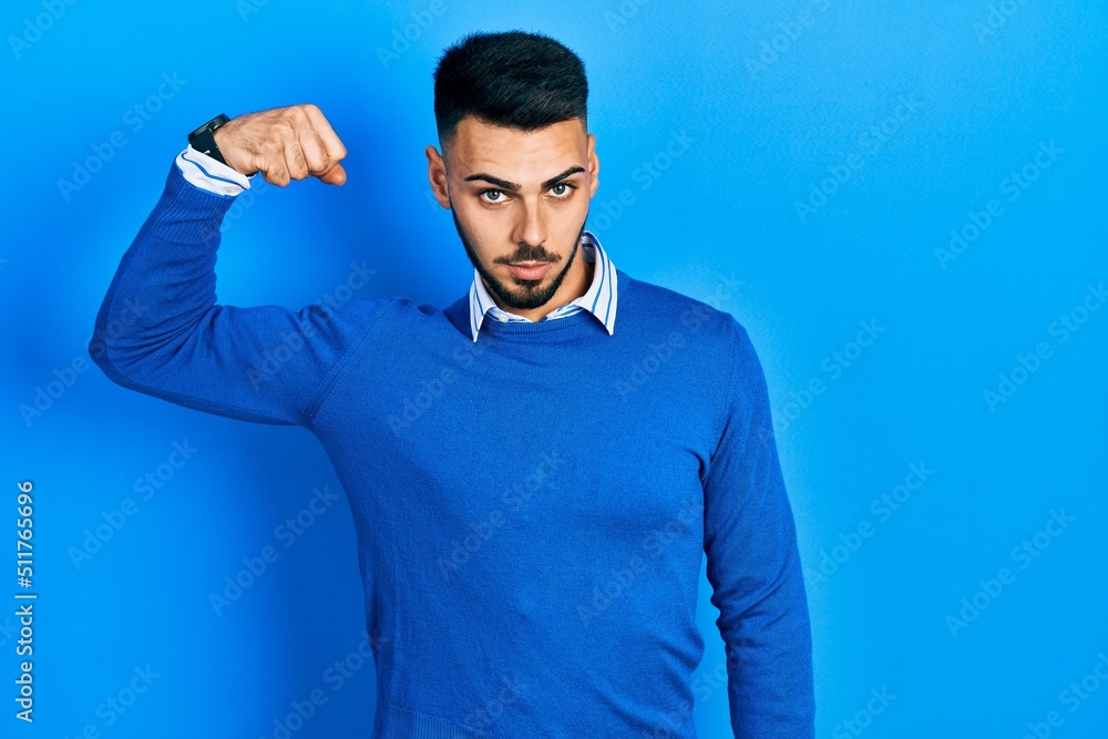 Naklejka premium Young hispanic man with beard wearing casual blue sweater strong person showing arm muscle, confident and proud of power