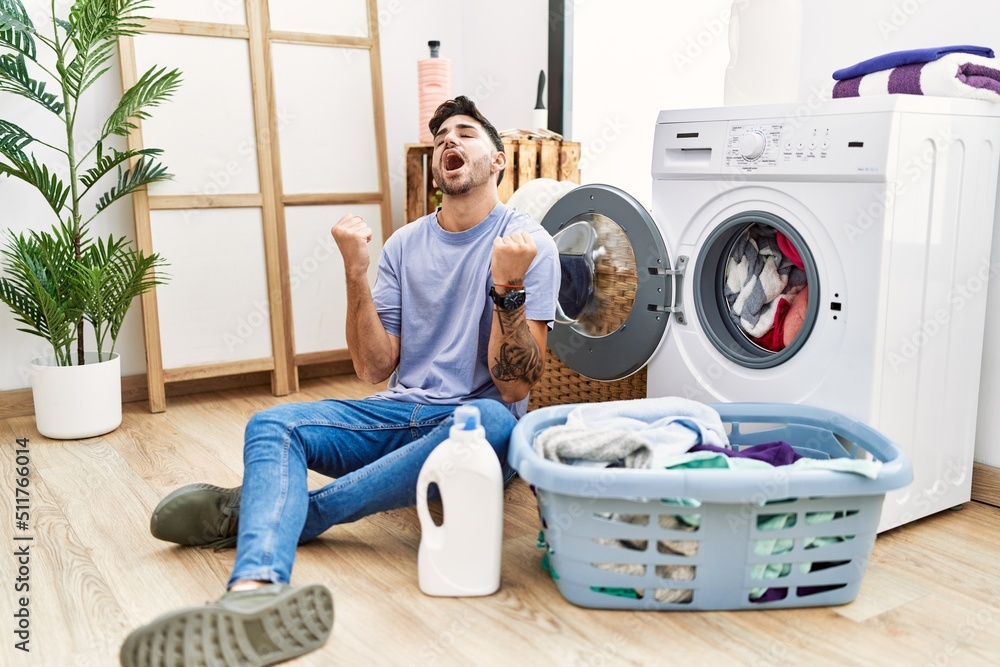Young hispanic man putting dirty laundry into washing machine celebrating surprised and amazed for success with arms raised and eyes closed. winner concept. Young hispanic man putting dirty laundry into washing machine celebrating surprised and amazed for success with arms raised and eyes closed. winner concept.