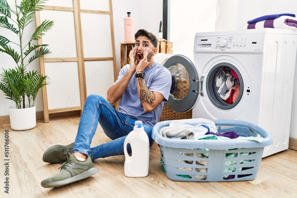 Young hispanic man putting dirty laundry into washing machine afraid and shocked, surprise and amazed expression with hands on face Young hispanic man putting dirty laundry into washing machine afraid and shocked, surprise and amazed expression with hands on face