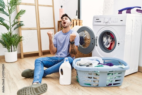 Young hispanic man putting dirty laundry into washing machine celebrating surprised and amazed for success with arms raised and eyes closed. winner concept.