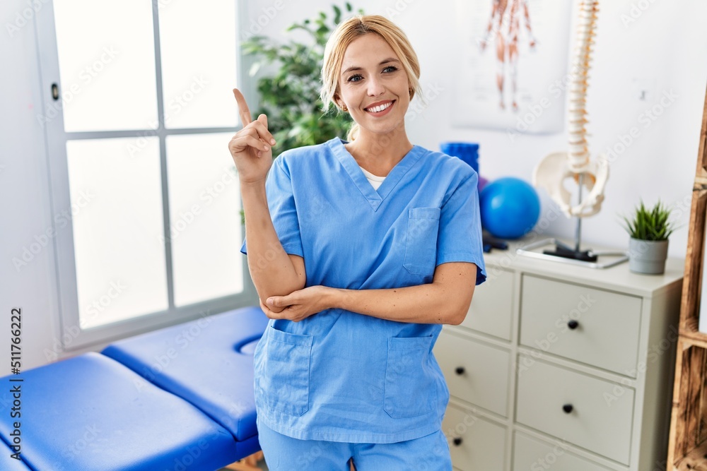 Beautiful blonde physiotherapist woman working at pain recovery clinic with a big smile on face, pointing with hand and finger to the side looking at the camera.