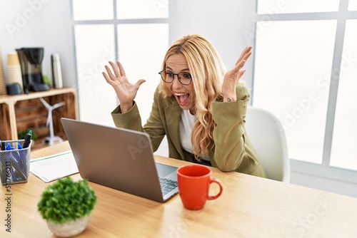 Beautiful blonde woman working at the office with laptop celebrating crazy and amazed for success with arms raised and open eyes screaming excited. winner concept