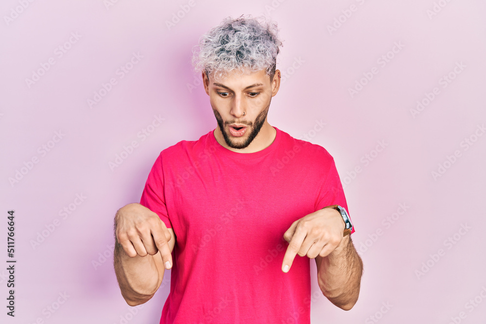 Young hispanic man with modern dyed hair wearing casual pink t shirt pointing down with fingers showing advertisement, surprised face and open mouth Young hispanic man with modern dyed hair wearing casual pink t shirt pointing down with fingers showing advertisement, surprised face and open mouth