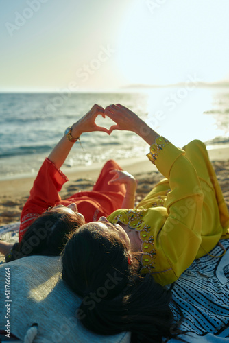 Two Arab women, lying on the beach, hugging smiling, wearing typical Moroccan clothes. Enjoying the summer, making a heart with their hand. Vertical photo