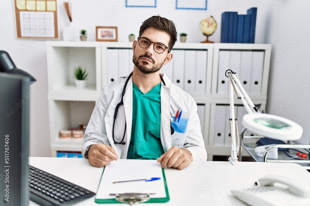 Young man with beard wearing doctor uniform and stethoscope at the clinic relaxed with serious expression on face. simple and natural looking at the camera.