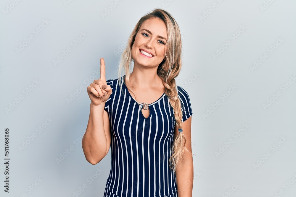 Beautiful young blonde woman wearing casual striped dress showing and pointing up with finger number one while smiling confident and happy.