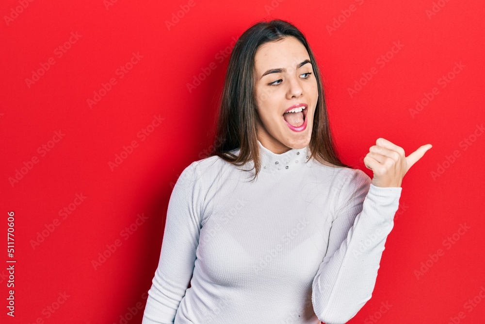 Young brunette teenager wearing white sweater pointing thumb up to the side smiling happy with open mouth Young brunette teenager wearing white sweater pointing thumb up to the side smiling happy with open mouth