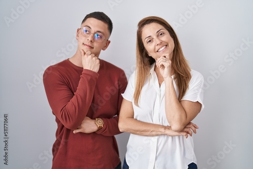 Mother and son standing together over isolated background with hand on chin thinking about question, pensive expression. smiling with thoughtful face. doubt concept.