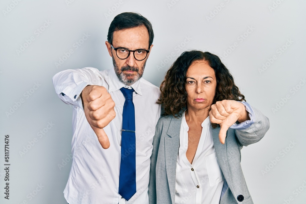 Middle age couple of hispanic woman and man wearing business office uniform looking unhappy and angry showing rejection and negative with thumbs down gesture. bad expression. Middle age couple of hispanic woman and man wearing business office uniform looking unhappy and angry showing rejection and negative with thumbs down gesture. bad expression.