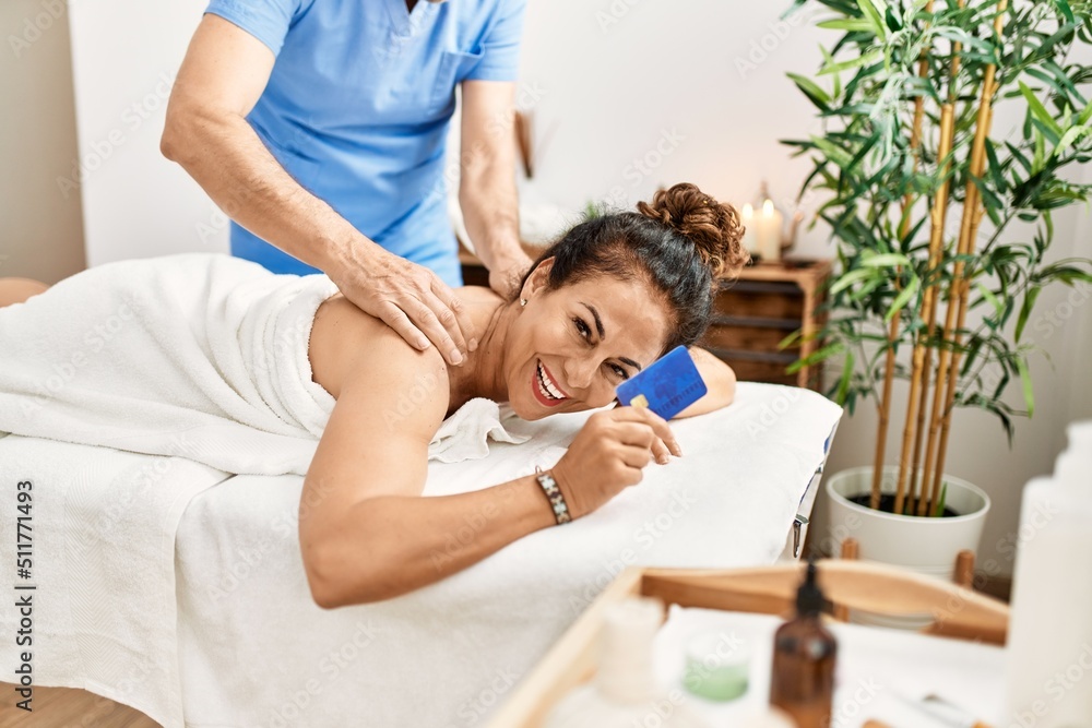 Middle age man and woman wearing therapist uniform having back massage session and holding credit card at beauty center Middle age man and woman wearing therapist uniform having back massage session and holding credit card at beauty center