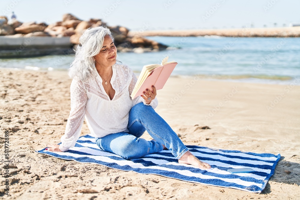 Middle age woman reading book sitting on towel at seaside