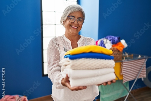 Middle age woman smiling confident holding folded clothes at laundry room