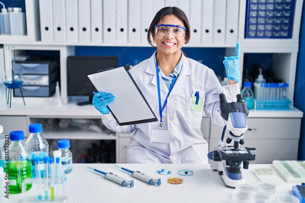 Hispanic young woman working at scientist laboratory smiling amazed and surprised and pointing up with fingers and raised arms. Hispanic young woman working at scientist laboratory smiling amazed and surprised and pointing up with fingers and raised arms.