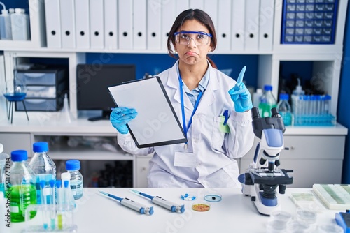 Hispanic young woman working at scientist laboratory pointing up looking sad and upset  indicating direction with fingers  unhappy and depressed.