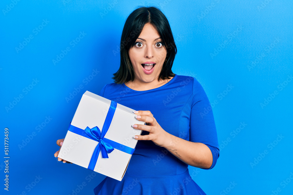 Young hispanic woman holding gift afraid and shocked with surprise and amazed expression, fear and excited face. Young hispanic woman holding gift afraid and shocked with surprise and amazed expression, fear and excited face.