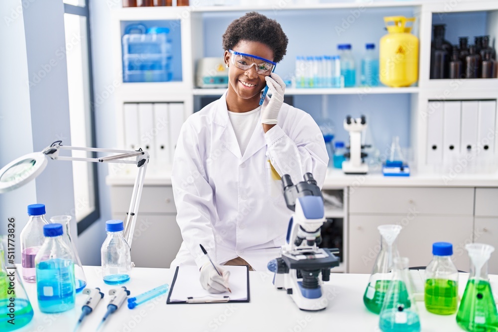 African american woman wearing scientist uniform talking on the smartphone write on clipboard at laboratory African american woman wearing scientist uniform talking on the smartphone write on clipboard at laboratory