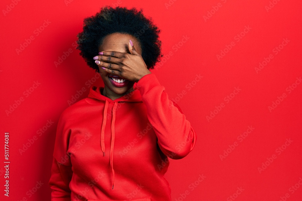 Young african american woman wearing casual sweatshirt smiling and laughing with hand on face covering eyes for surprise. blind concept. Young african american woman wearing casual sweatshirt smiling and laughing with hand on face covering eyes for surprise. blind concept.