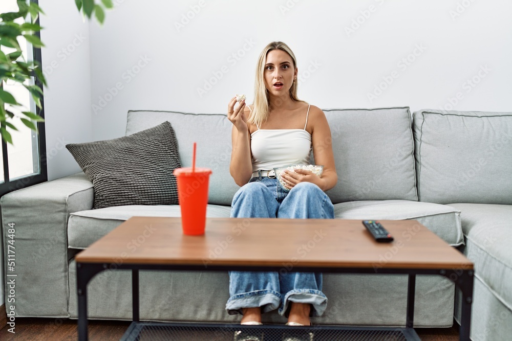 Young blonde woman eating popcorn sitting on the sofa scared and amazed with open mouth for surprise, disbelief face