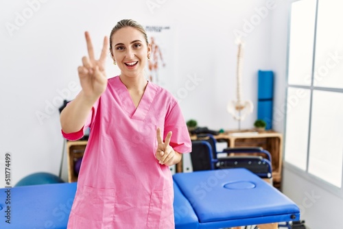 Young blonde woman working at pain recovery clinic smiling looking to the camera showing fingers doing victory sign. number two.