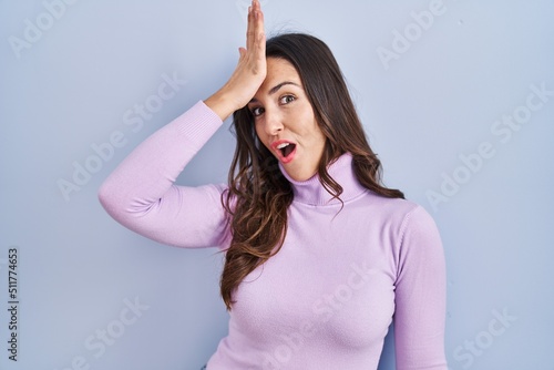 Young brunette woman standing over blue background surprised with hand on head for mistake, remember error. forgot, bad memory concept.