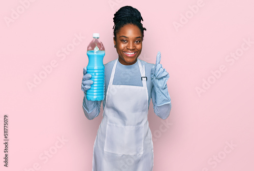 Young african american woman wearing apron holding detergent bottle surprised with an idea or question pointing finger with happy face, number one