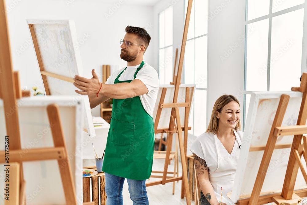 Obraz premium Young caucasian couple smiling happy drawing at art studio. Man standing and holding canvas.
