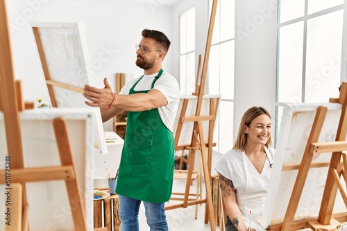 Young caucasian couple smiling happy drawing at art studio. Man standing and holding canvas.