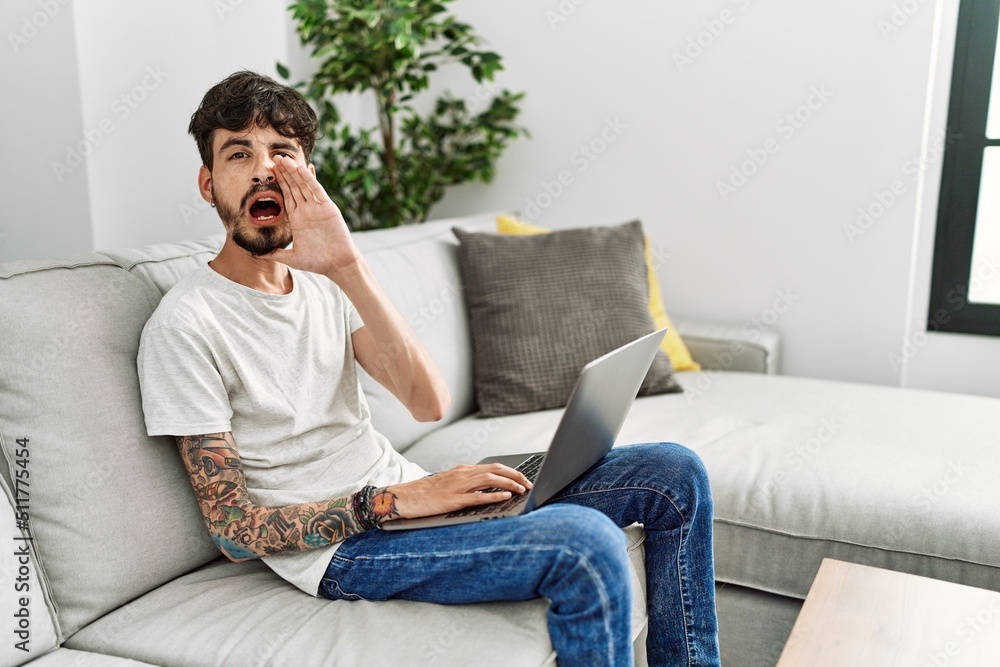 Hispanic man with beard sitting on the sofa shouting and screaming loud to side with hand on mouth. communication concept.