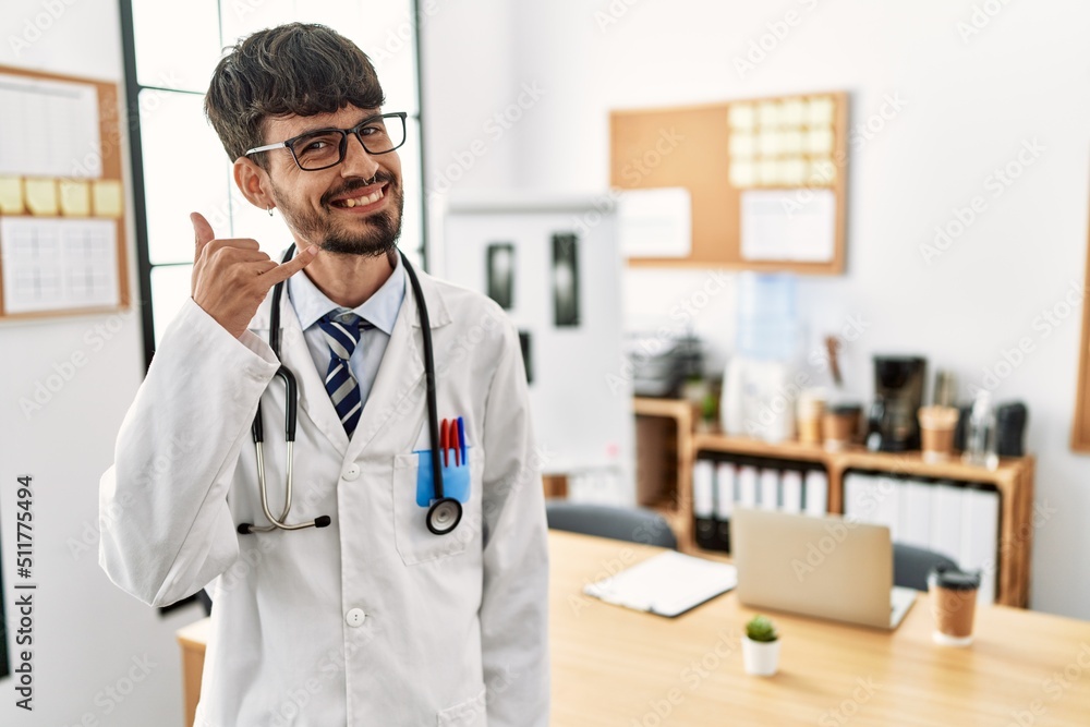Hispanic man with beard wearing doctor uniform and stethoscope at the office smiling doing phone gesture with hand and fingers like talking on the telephone. communicating concepts. Hispanic man with beard wearing doctor uniform and stethoscope at the office smiling doing phone gesture with hand and fingers like talking on the telephone. communicating concepts.
