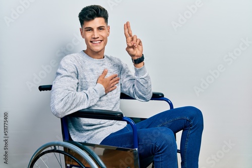 Young hispanic man sitting on wheelchair smiling swearing with hand on chest and fingers up, making a loyalty promise oath