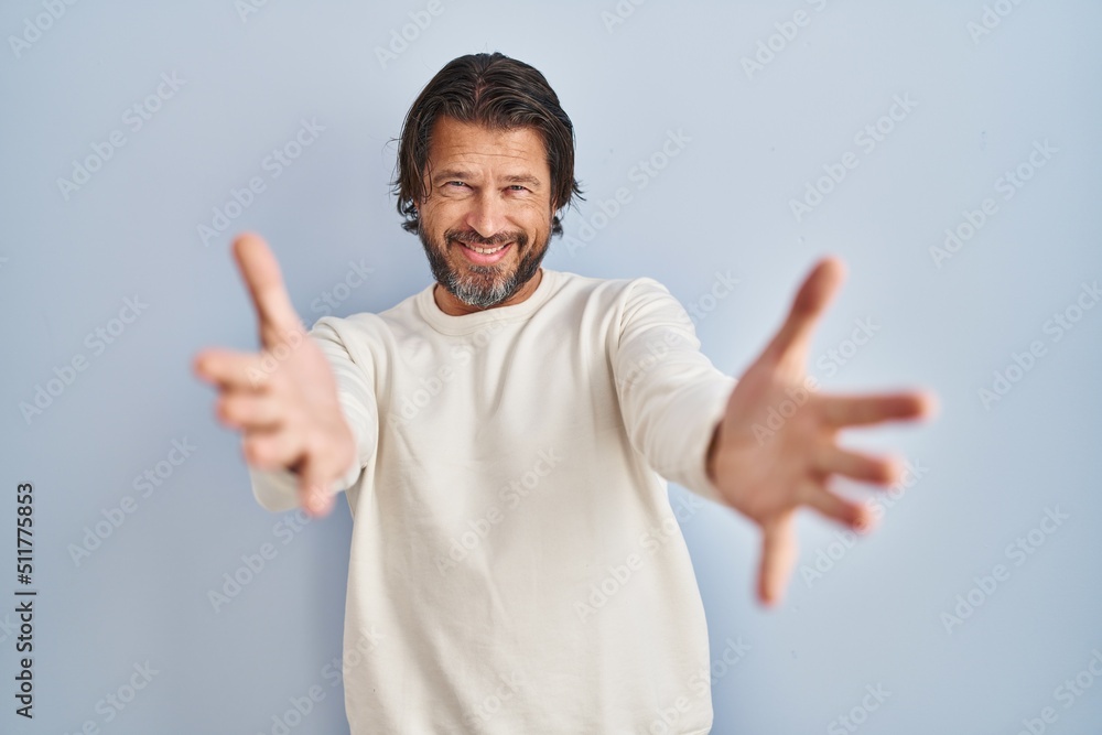 Fototapeta premium Handsome middle age man wearing casual sweater over blue background looking at the camera smiling with open arms for hug. cheerful expression embracing happiness.