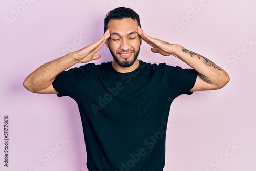 Hispanic man with beard wearing casual black t shirt with hand on head, headache because stress. suffering migraine.