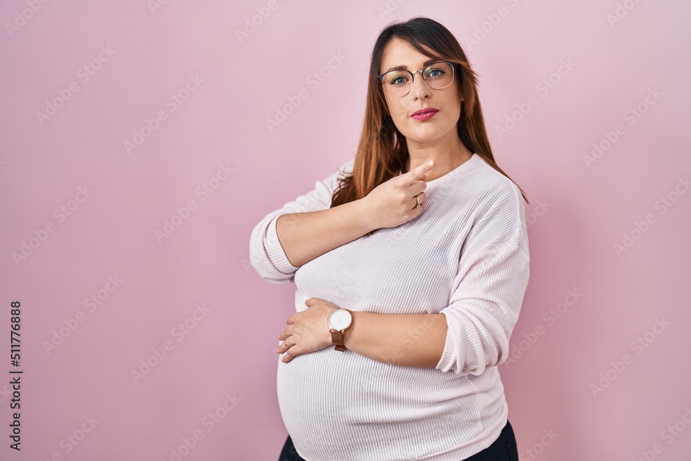 Fototapeta premium Pregnant woman standing over pink background pointing with hand finger to the side showing advertisement, serious and calm face