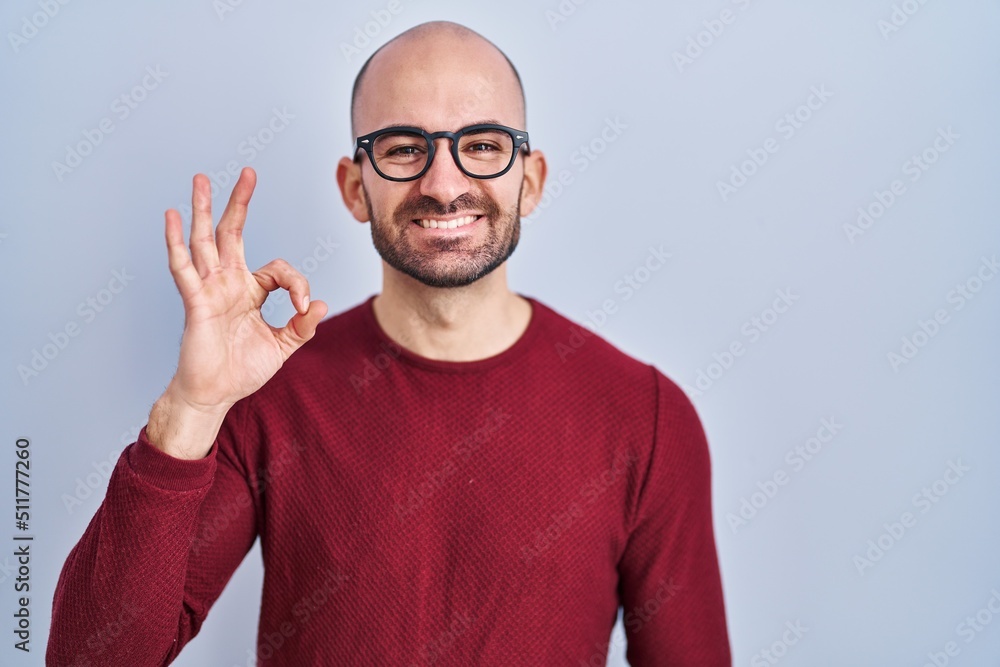 Young bald man with beard standing over white background wearing glasses smiling positive doing ok sign with hand and fingers. successful expression. Young bald man with beard standing over white background wearing glasses smiling positive doing ok sign with hand and fingers. successful expression.
