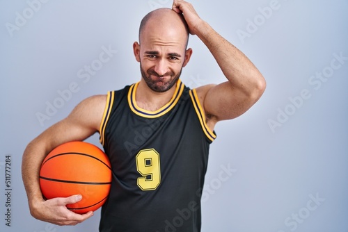 Young bald man with beard wearing basketball uniform holding ball confuse and wondering about question. uncertain with doubt, thinking with hand on head. pensive concept.