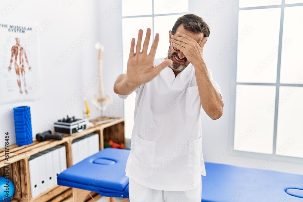 Middle age man with beard working at pain recovery clinic covering eyes with hands and doing stop gesture with sad and fear expression. embarrassed and negative concept. Middle age man with beard working at pain recovery clinic covering eyes with hands and doing stop gesture with sad and fear expression. embarrassed and negative concept.