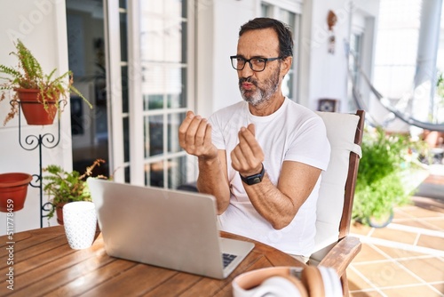 Middle age man using computer laptop at home doing money gesture with hands, asking for salary payment, millionaire business photo