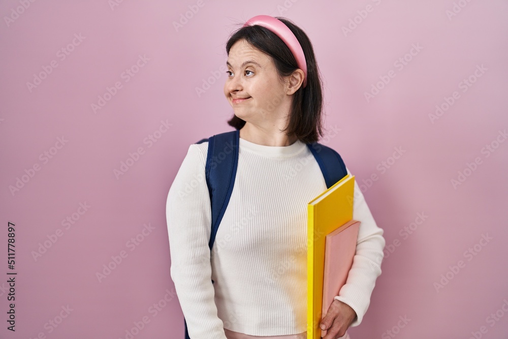 Woman with down syndrome wearing student backpack and holding books looking away to side with smile on face, natural expression. laughing confident.