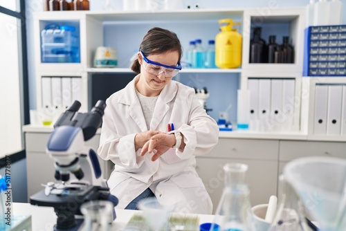 Hispanic girl with down syndrome working at scientist laboratory in hurry pointing to watch time  impatience  upset and angry for deadline delay