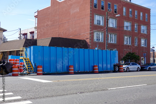 A large blue dumpster on a street near a large brick building with orange reflective warning barrels