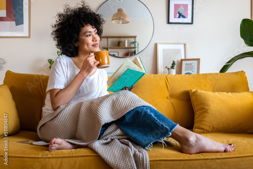 Fotografia Pensive relaxed African american woman reading a book at home, drinking coffee sitting on the couch