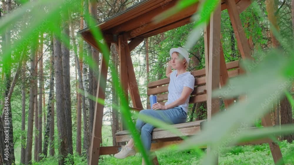 Romantic adult woman sitting on wooden swing in beautiful park among trees. View through green leaves, focus on the background. Summer lifestyle, harmony with nature concept.