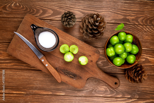 top view of sliced green plums with salt and kitchen knife on a wooden cutting board , cones and plums in a bowl on rustic wooden background