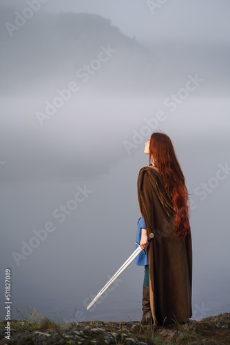 Red-haired girl in armor with sword and raincoat on river bank