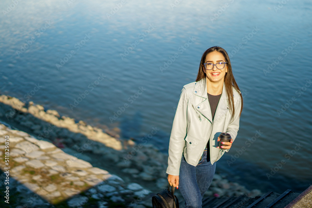 Beautiful young woman walking near river in city while holding coffee cup and her bag. Beautiful young woman walking near river in city while holding coffee cup and her bag.