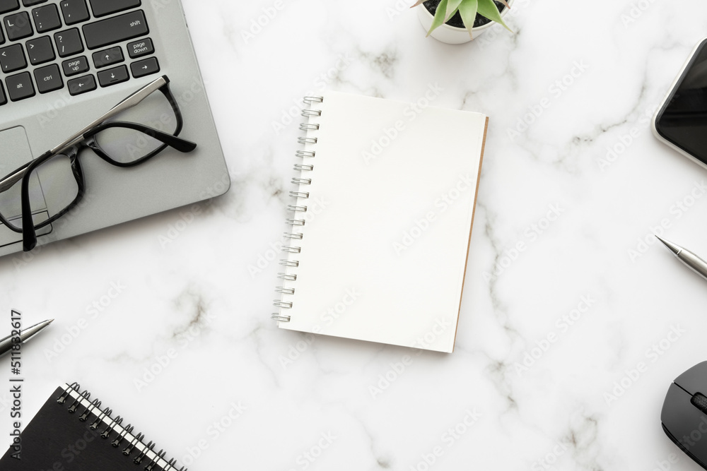 Blank notebook is on top of white marble office desk table with laptop computer and office supplies. Top view with copy space, flat lay. Blank notebook is on top of white marble office desk table with laptop computer and office supplies. Top view with copy space, flat lay.