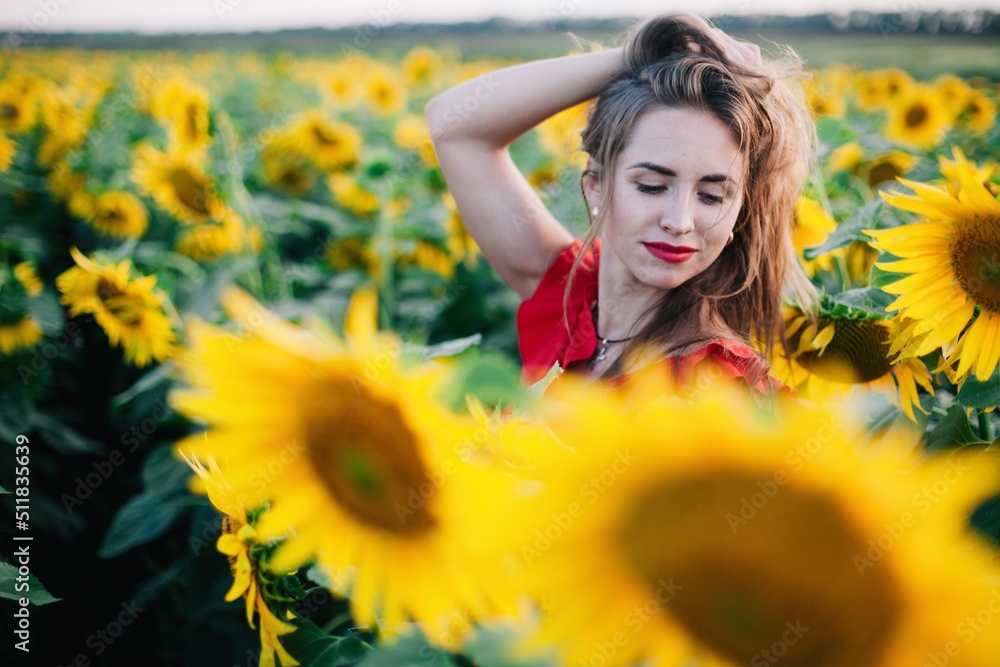 A young, slender girl with loose hair in a red dress and straw hat stands in a field of sunflowers at sunset A young, slender girl with loose hair in a red dress and straw hat stands in a field of sunflowers at sunset