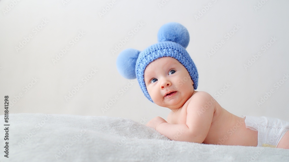 A smiling infant baby in a blue hat with pompoms looks into the camera. A happy newborn is lying on a blanket. Beautiful, happy little child with blue eyes. Cheerful White kid. A smiling infant baby in a blue hat with pompoms looks into the camera. A happy newborn is lying on a blanket. Beautiful, happy little child with blue eyes. Cheerful White kid.