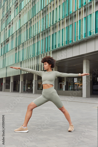 Full length shot of motivated curly haired sporty woman stretches arms exercises outdoors dressed in activewear poses near modern urban building focused into distance. Workout and sport concept