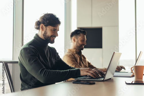 Joyful male coworkers using laptops, working on business project together as team in company office, blank space